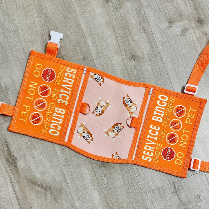 An orange and pink service dog vest with printed fabric featuring Bingo from the cartoon Bluey. Text on the side panels reads: "Service Bingo, cheese and crackers do not pet"