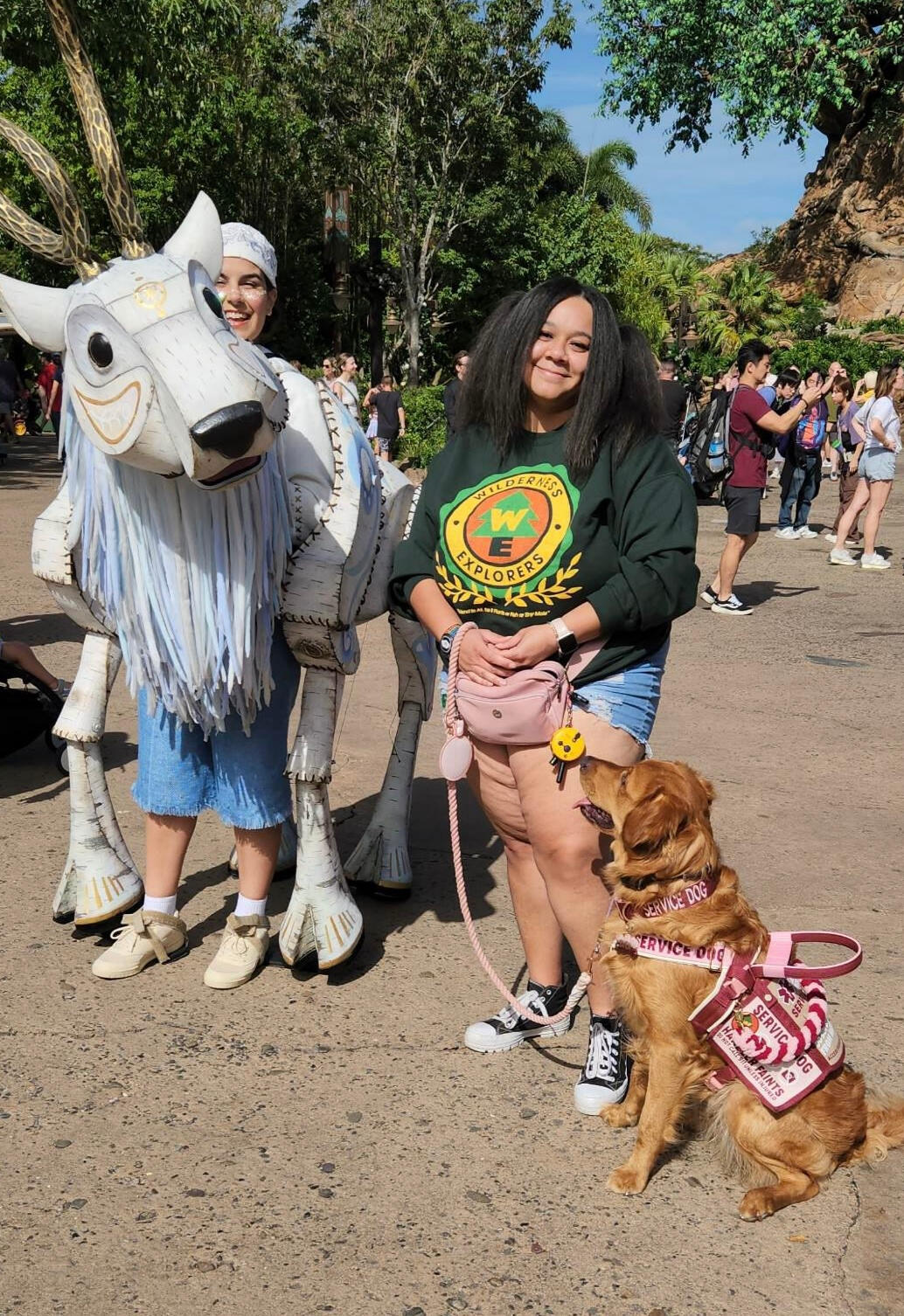 A plus size black woman with long dark brown hair looking at the camera and smiling while wearing a pink dress. She is also holding a rollator mobility aide, and is pictured standing in a botanical garden.