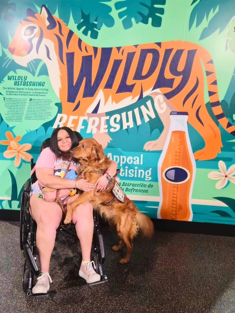 A plus size black woman with long dark brown hair looking at the camera and smiling while wearing a pink dress. She is also holding a rollator mobility aide, and is pictured standing in a botanical garden.